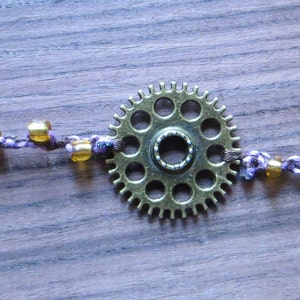 May include: A steampunk-style bracelet featuring a central bronze gear, connected by a purple and white braided cord. The cord is adorned with small, golden beads. The bracelet has a bronze clasp.