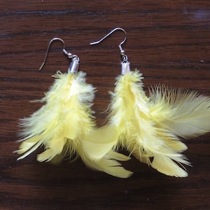 Può includere: Un paio di orecchini pendenti con piume giallo brillante. Gli orecchini hanno ganci color argento e piccoli dettagli metallici. Le piume sono morbide e soffici, con un aspetto naturale e strutturato. Gli orecchini sono esposti su una superficie di legno marrone scuro.