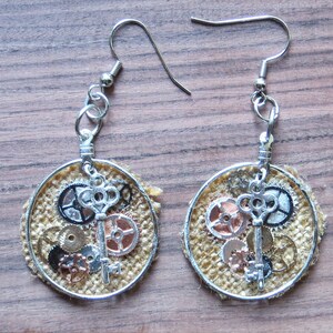 Può includere: Orecchini in stile steampunk con un design circolare. Presentano un supporto in tela di iuta, una cornice argentata e piccoli ingranaggi in argento, rame e nero. Un ciondolo a forma di chiave argentata è appeso al centro.
