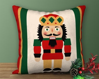 May include: Square decorative pillow featuring a colorful illustration of a nutcracker soldier. The pillow has a cream-colored center with a red, green, and gold border. The nutcracker is dressed in a red uniform with a black beard and a gold crown.