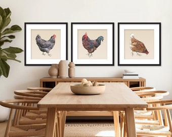 May include: Three framed illustrations of chickens, each in a black frame with a white mat. The illustrations are of a black and white hen, a brown rooster, and a brown hen. The illustrations are set against a white wall and a wooden table with chairs.