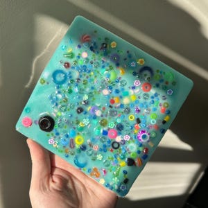 May include: A square teal blue resin coaster with a variety of colorful beads and small trinkets embedded in the surface.