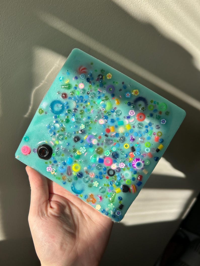 May include: A square teal blue resin coaster with a variety of colorful beads and small trinkets embedded in the surface.