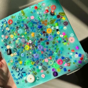May include: A square, teal blue resin tray filled with colorful, assorted beads, charms, and glitter. The tray is translucent, allowing the light to shine through the beads.
