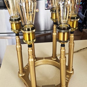 May include: A gold-colored, multi-bulb lamp with Edison-style light bulbs. The lamp has a hexagonal base and cylindrical supports. The light bulbs are clear with visible filaments. The lamp has a switch box with three buttons.