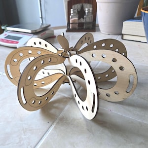 May include: A wooden, geometric sculpture with a butterfly-like design. The sculpture is made of light-colored wood and features multiple curved, openwork sections with evenly spaced holes. The pieces are interlocked to create a three-dimensional form.