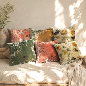 Puede incluir: Seis cojines cuadrados dispuestos sobre un diván. Los cojines presentan texturas y colores variados, incluyendo superficies lisas de aspecto aterciopelado en verde oscuro, naranja quemado y amarillo mostaza, junto a diseños florales.