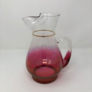 May include: A clear glass pitcher with a pink ombre design and a gold band around the top. The pitcher has a handle and a wide mouth.