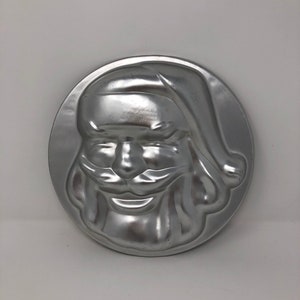 May include: A silver metal mold shaped like Santa Claus's face. The mold is designed for baking and has a detailed design of Santa's face, beard, and hat.