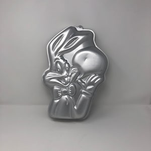 May include: A silver metal cake pan shaped like Bugs Bunny. The pan is designed to create a cake that looks like Bugs Bunny's head and shoulders.