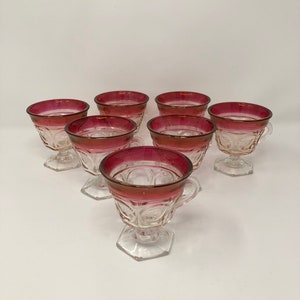May include: Set of seven clear glass cups with a red rim. The cups have a pedestal base and a handle.