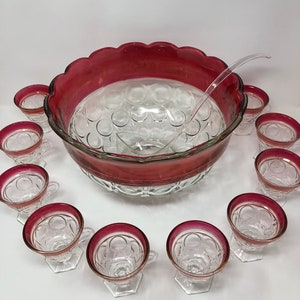 May include: A vintage glass punch bowl set with a red rim and clear glass cups. The bowl has a scalloped edge and a textured design. The set includes a ladle.
