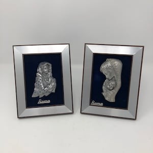 May include: Two framed silver-toned relief sculptures. Each frame has a silver border and a dark blue background. One depicts a woman holding a child, the other a woman with a headscarf holding a child. The word "Roma" is at the bottom.