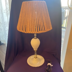 May include: A vintage table lamp with a tan rope shade and a white hobnail base. The lamp has a brass-colored stem and a scalloped base. The power cord is brown and wrapped with a white tie.