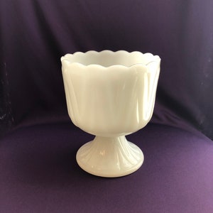 May include: A white milk glass pedestal vase with a scalloped rim and a fluted design. The vase is on a purple background.