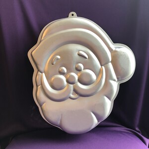 May include: A silver metal Santa Claus cake pan with a smiling face, a white beard, and a red hat.