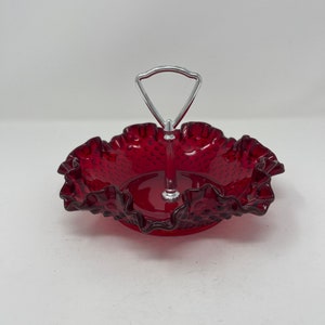 May include: A red glass candy dish with a ruffled edge and a silver handle. The dish has a textured surface and is perfect for serving small treats.