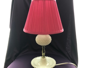 May include: A white milk glass lamp with a pink pleated lampshade. The lamp has a round base with a beaded edge and a tall, slender stem.
