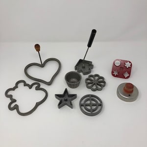 May include: A set of vintage metal cookie cutters in various shapes, including a heart, a star, a flower, a circle, a house, and a cloud. There is also a red plastic cookie cutter with various shapes and a metal cookie cutter with a wooden handle.