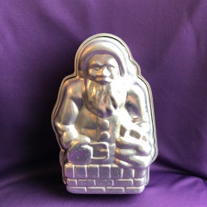 May include: A silver metal Santa Claus cake pan. The pan is shaped like Santa Claus sitting in a chimney.