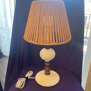May include: A white milk glass table lamp with a brown rope shade. The lamp has a round base and a wooden stem.