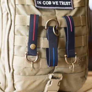 Puede incluir: Tres llaveros de cuero con rayas de diferentes colores, unidos a una mochila t&aacute;ctica beige. Los llaveros tienen anillas en D de lat&oacute;n y est&aacute;n unidos a la mochila con un clip de metal.