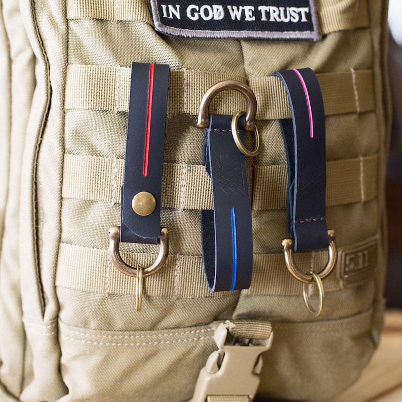 Puede incluir: Tres llaveros de cuero con rayas de diferentes colores, unidos a una mochila t&aacute;ctica beige. Los llaveros tienen anillas en D de lat&oacute;n y est&aacute;n unidos a la mochila con un clip de metal.