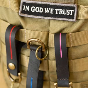 Puede incluir: Primer plano de una bolsa de lona marr&oacute;n con dos correas de cuero. Las correas tienen costuras rojas y rosadas. La bolsa tiene un anillo de lat&oacute;n y un parche que dice "In God We Trust".