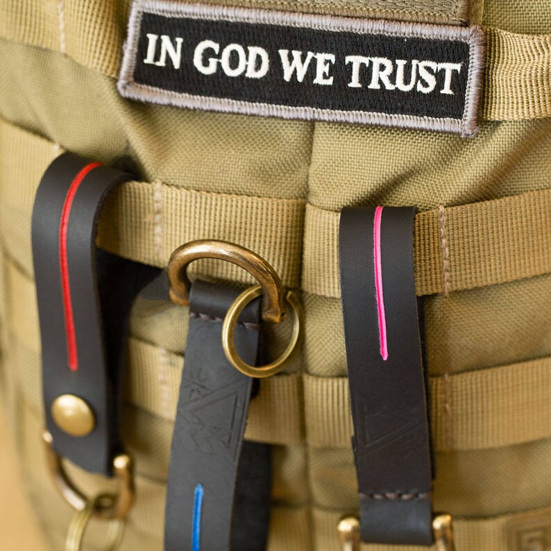 Puede incluir: Primer plano de una bolsa de lona marr&oacute;n con dos correas de cuero. Las correas tienen costuras rojas y rosadas. La bolsa tiene un anillo de lat&oacute;n y un parche que dice "In God We Trust".