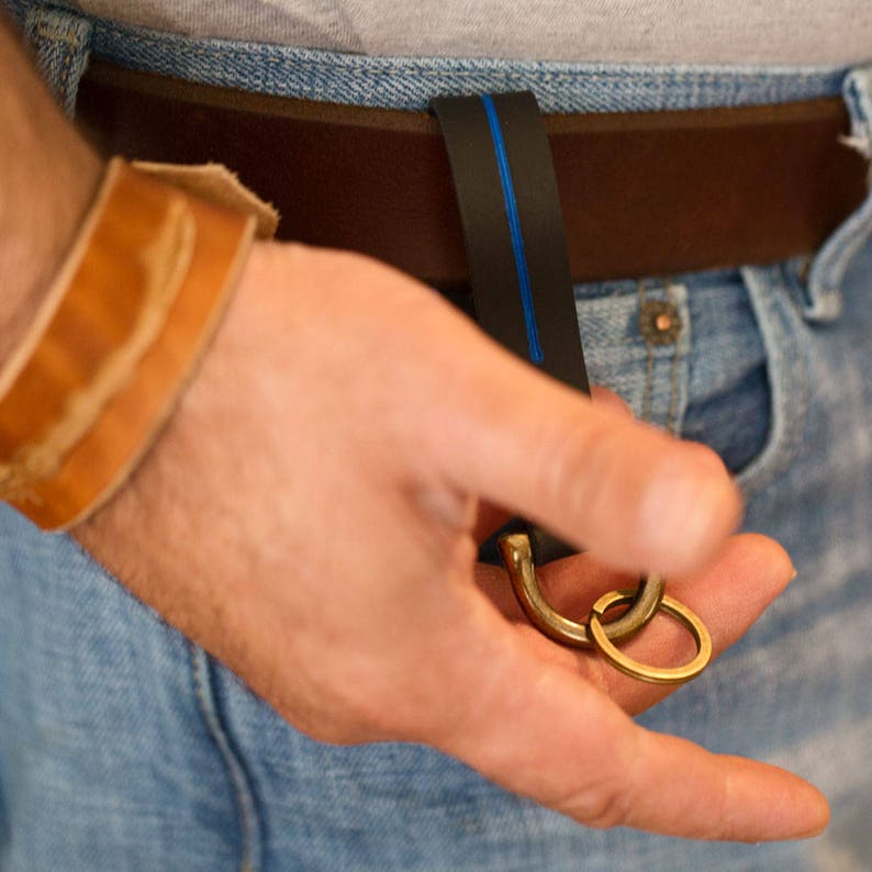 Puede incluir: Una persona que lleva pantalones vaqueros azules y un cintur&oacute;n de cuero marr&oacute;n con una raya azul. El cintur&oacute;n tiene un llavero de lat&oacute;n.