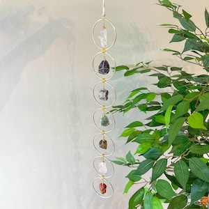 May include: A hanging decorative piece featuring seven gold-colored metal rings, each holding a different colored raw crystal. The crystals include clear, purple, gray, green, brown, pink, and red stones. A white string is used for hanging.