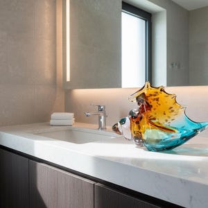 May include: A decorative glass seashell sculpture in shades of yellow, orange, and blue sits on a white countertop in a modern bathroom. The sculpture is near a sink, tap, and a stack of white towels.