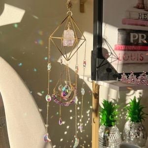 May include: A gold-colored geometric suncatcher with a raw amethyst crystal and iridescent crystal accents. The suncatcher features a diamond-shaped frame, dangling chains, and crescent moon charms. It is designed to catch sunlight and create a rainbow effect.