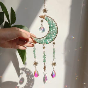 Puede incluir: Un atrapasoles en forma de media luna con un marco de metal dorado lleno de piedras verdes. El atrapasoles tiene hebras colgantes de piedras verdes, detalles en forma de estrella y colgantes de cristal iridiscente. Para la decoración del hogar.