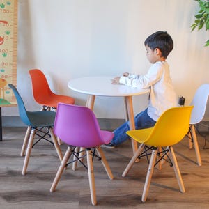 Op de afbeelding: Een witte tafel met vier stoelen, waarvan één geel, één paars, één teal en één oranje. Een jong persoon zit aan de tafel, leunend tegen de gele stoel.