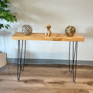 Può includere: Una consolle in legno chiaro con gambe a forcina nere. Il tavolo è decorato con una piccola statuina di cucciolo e due sfere decorative in metallo. Il tavolo è posto contro un muro bianco.