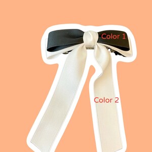 Pode incluir: Um la&ccedil;o de cabelo preto e branco com um la&ccedil;o grande no topo e duas caudas longas. O la&ccedil;o est&aacute; etiquetado como "Cor 1" e as caudas est&atilde;o etiquetadas como "Cor 2".