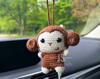 May include: A brown and white crocheted monkey hanging from a rearview mirror in a car. The monkey has black eyes and a small smile. The text "JS Boutique Gifts" is visible on the monkey's chest.