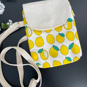 May include: A small white canvas crossbody bag with a lemon print. The bag has a flap closure and a long adjustable strap.