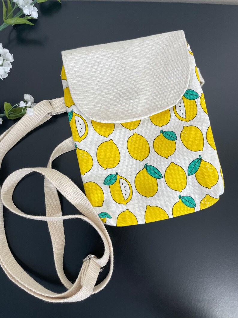 May include: A small white canvas crossbody bag with a lemon print. The bag has a flap closure and a long adjustable strap.