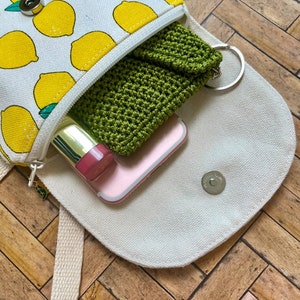 May include: A white canvas bag with a lemon print design. The bag is open and has a green crocheted pouch, a pink phone case, and a gold lipstick inside.
