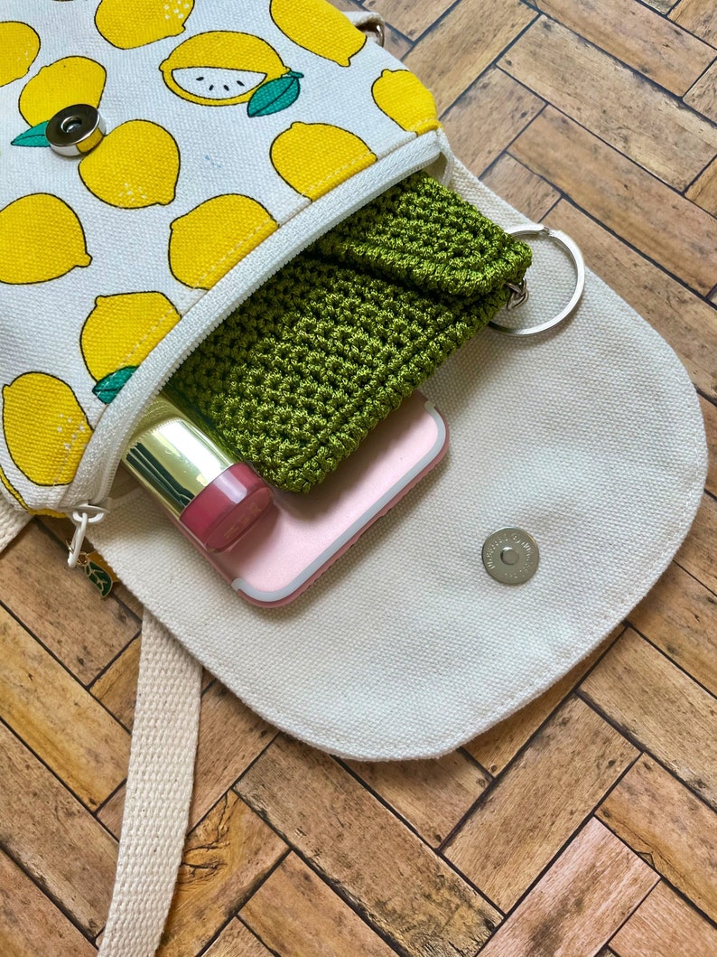 May include: A white canvas bag with a lemon print design. The bag is open and has a green crocheted pouch, a pink phone case, and a gold lipstick inside.
