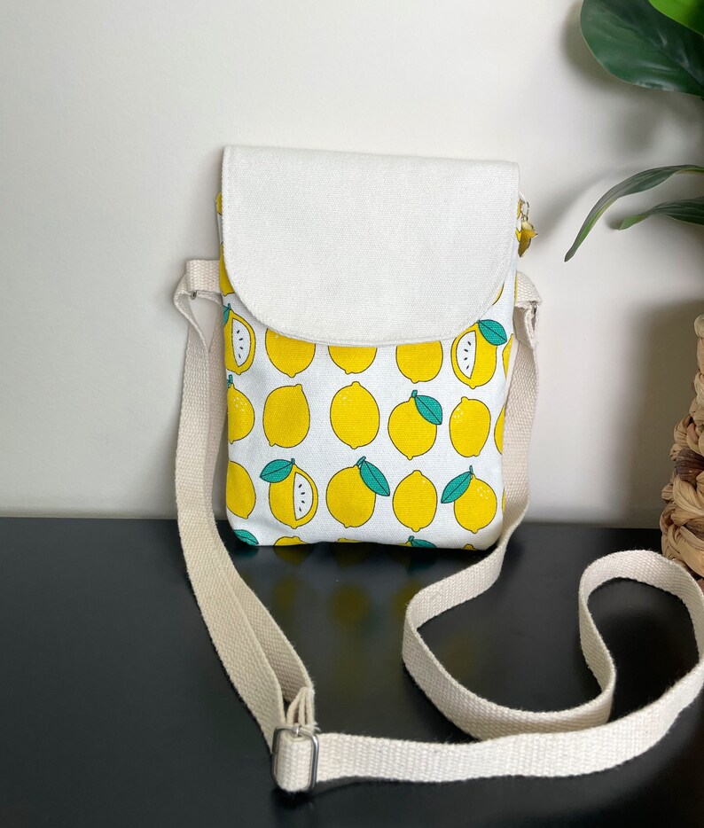May include: A small white canvas crossbody bag with a lemon print. The bag has a flap closure and an adjustable strap.