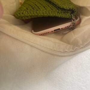 May include: A green crocheted pouch with a wooden button closure, a rose gold smartphone, and a silver key chain inside a white canvas bag.