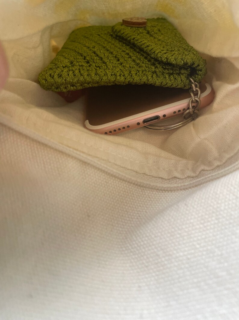 May include: A green crocheted pouch with a wooden button closure, a rose gold smartphone, and a silver key chain inside a white canvas bag.