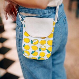 May include: A white canvas crossbody bag with a lemon print. The bag has a flap closure and a white strap.