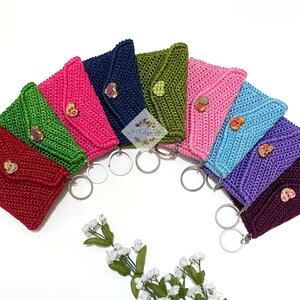 May include: A set of eight crocheted keychains in various colors, including red, green, pink, purple, and blue. Each keychain features a small, floral button and is attached to a silver key ring.