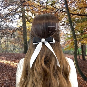 May include: A black and white satin ribbon bow hair clip. The bow is tied in a classic shape with long tails. The hair clip is worn in a woman's hair, with the background showing a blurred image of an autumn forest.