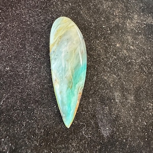 May include: A teardrop-shaped polished gemstone. The stone displays a mix of teal, light blue, and brown hues, with a glossy surface. The gemstone is set against a dark backdrop.