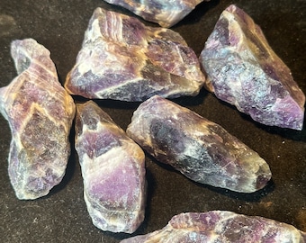 Puede incluir: Una colección de seis cristales de amatista, cada uno con una forma y un tamaño únicos. Los cristales son de color morado oscuro con vetas blancas y grises.